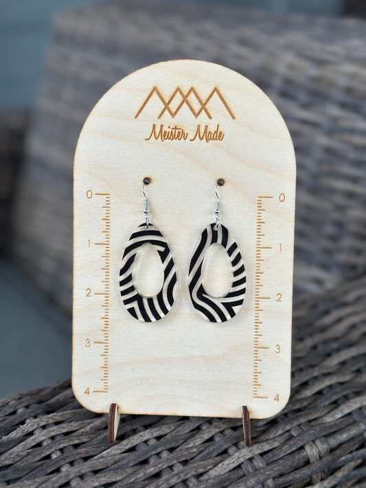 A pair of black and clear swirly acrylic earrings displayed on a wooden stand with the brand name 'Meister Made'.