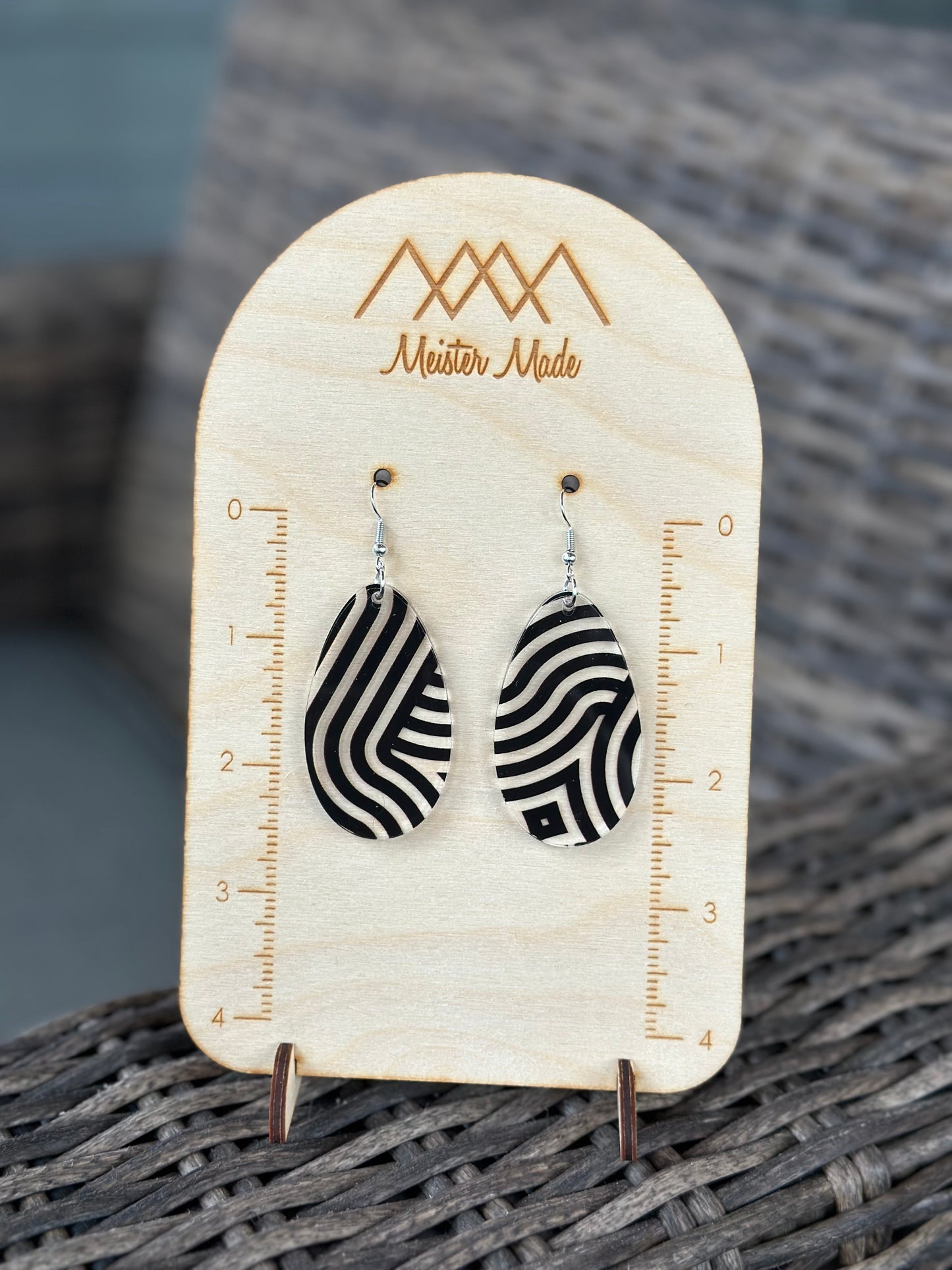 A pair of black and clear swirly acrylic earrings displayed on a wooden stand with the brand name 'Meister Made'.