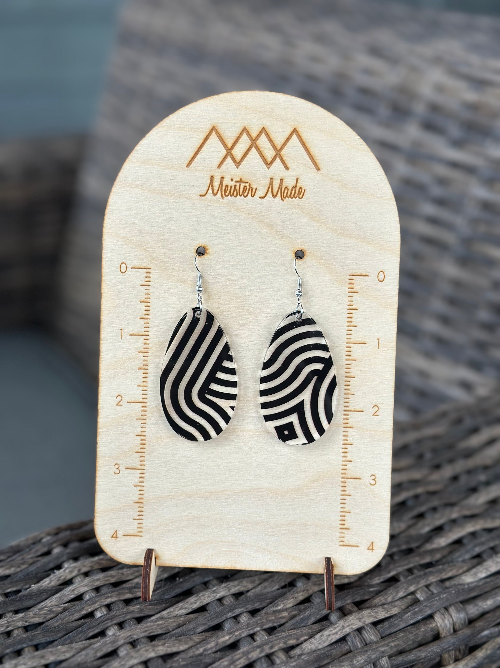 A pair of black and clear swirly acrylic earrings displayed on a wooden stand with the brand name 'Meister Made'.