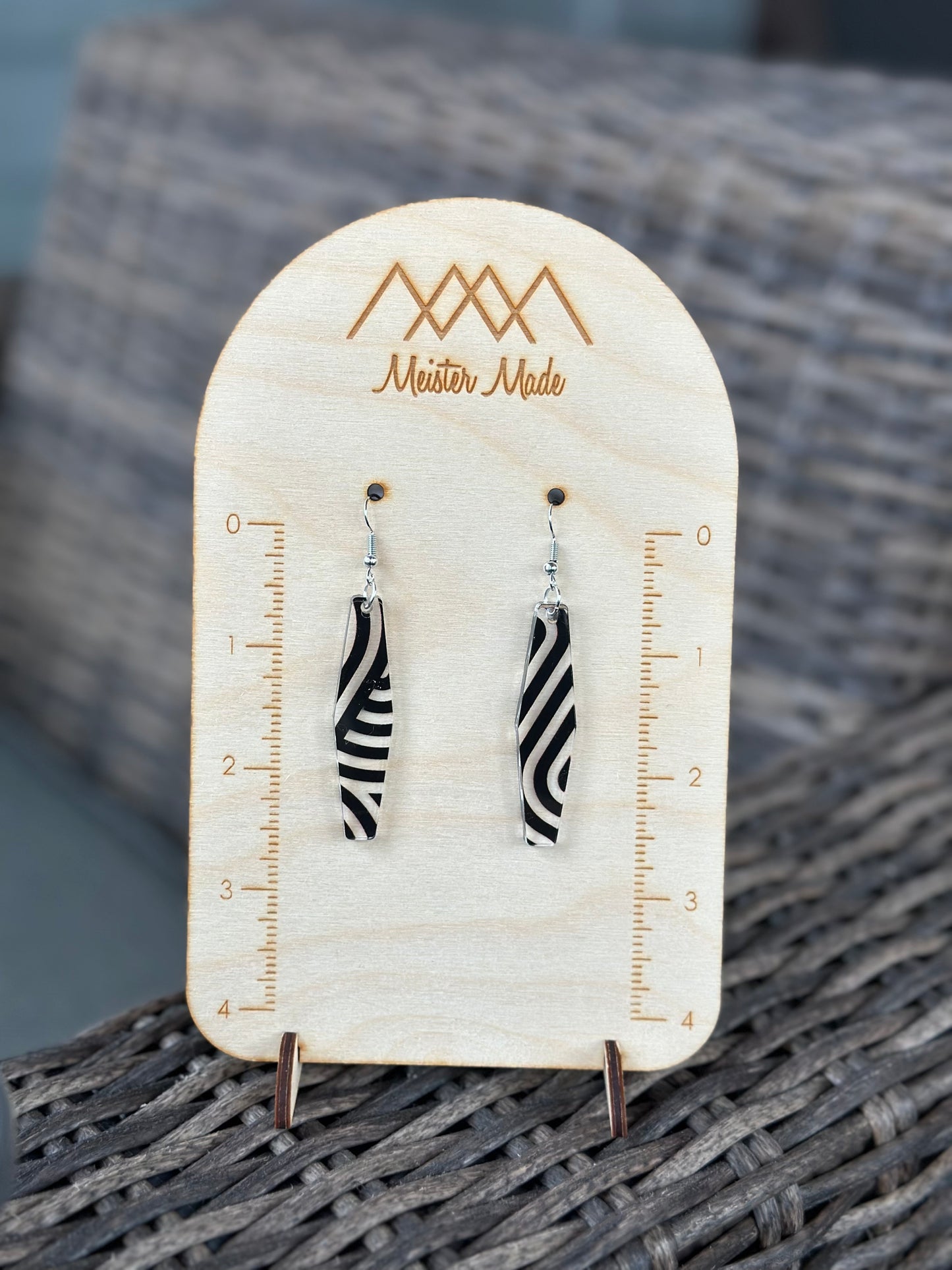 A pair of black and clear swirly acrylic earrings displayed on a wooden stand with the brand name 'Meister Made'.