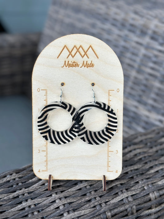 A pair of black and clear swirly acrylic earrings displayed on a wooden stand with the brand name 'Meister Made'.